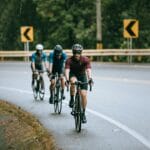 Dwóch rowerzystów w kaskach jedzie latem szosą oddzieloną barierkami
